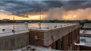 Fotografia do topo de um edifício comercial de tijolos e concreto, mostrando um sistema de proteção contra descargas atmosféricas (SPDA) instalado. Vários captores de cobre (para-raios) e cabos condutores percorrem a laje molhada e descem pela parede. O fundo é um céu dramático de tempestade durante o pôr do sol, com nuvens pesadas, cortinas de chuva e um raio caindo no horizonte.